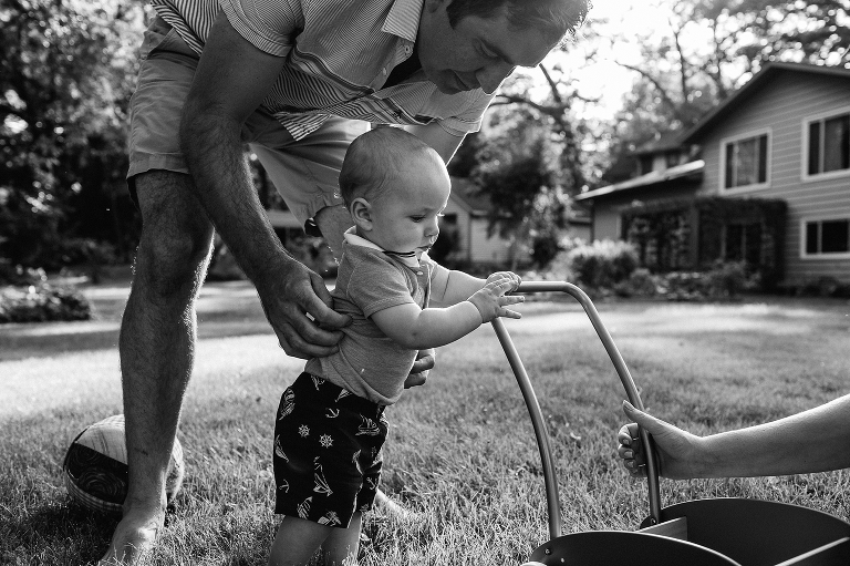 Black and white. Father holds up baby pushing a little cart learning how to walk.