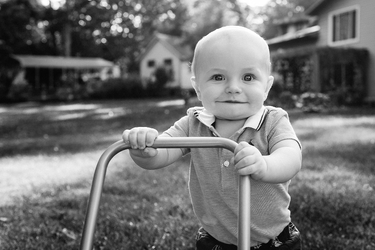 Black and white. Baby boy holds pushes baby toy to help him walk through the grass.