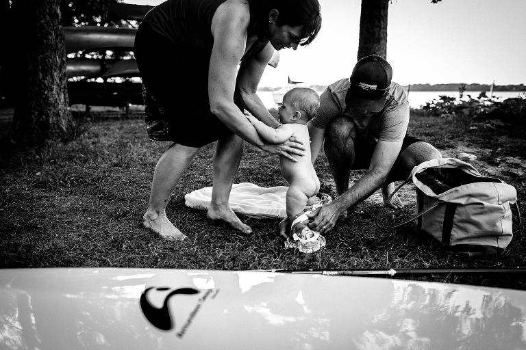 Black and white. Mother holds onto son as father pulls on baby pants changing him after going swimming in the lake.