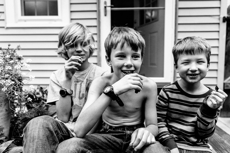 Black and white. Three boys sit outside house all eating bacon.