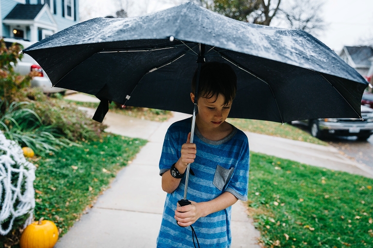 Boy stands outside in the rain holding black umbrella walking up the sidewalk. halloween decorations and fall leaves in the background.