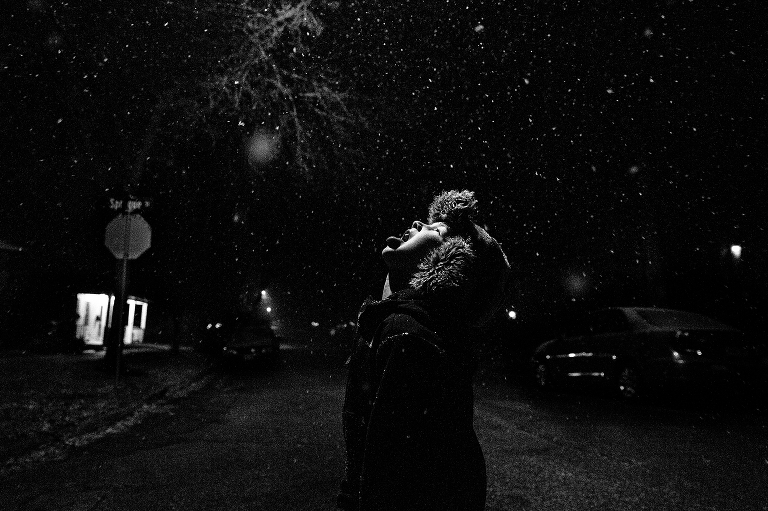 Black and white. Boy in street at night sticking tongue out to catch snowflakes. streetlight is source of light