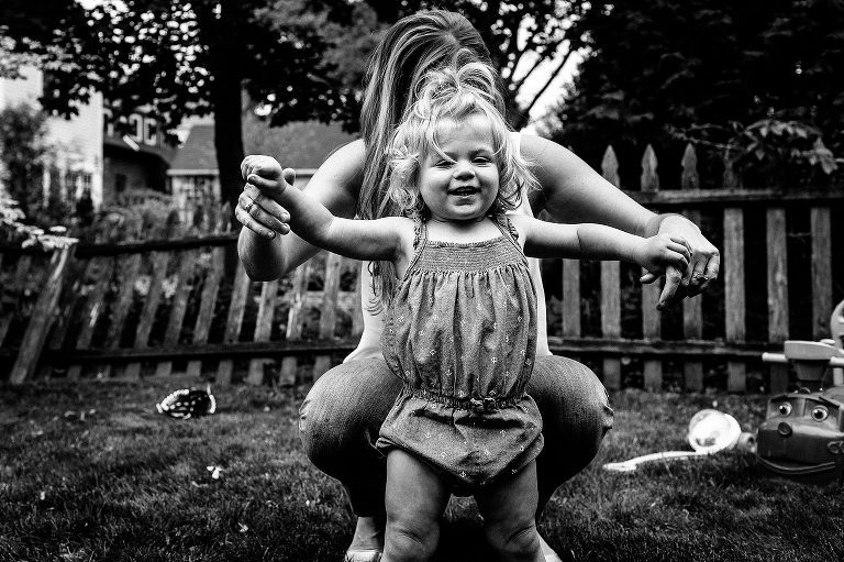 Black and white. Mother holds young daughters hands helping her walk around back yard.