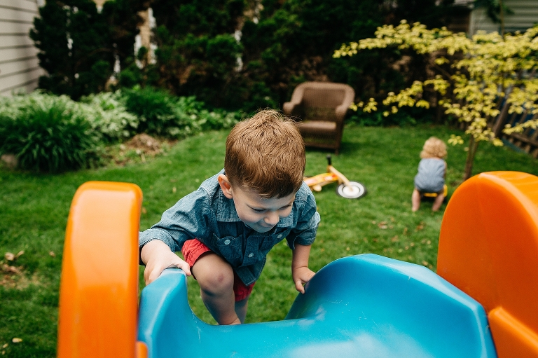 Young boy climbs up slide as younger sister plays in the grass.