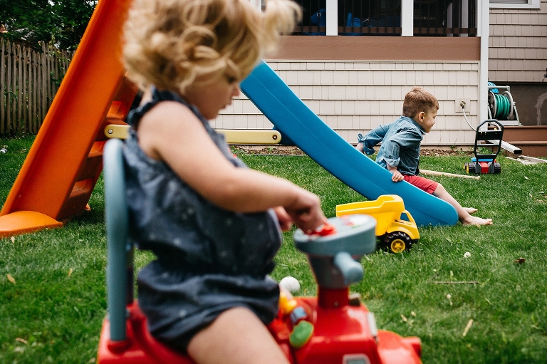 Young girl sitting on play toy as brother slides down a blue slide.