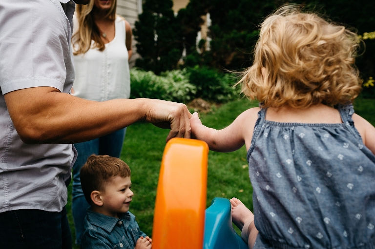 Mother and son watch father help his daughter down a slide.