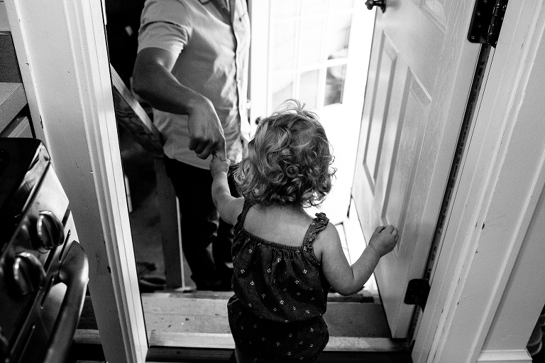 Black and white. Father helps young daughter walk outside the back door of the house.