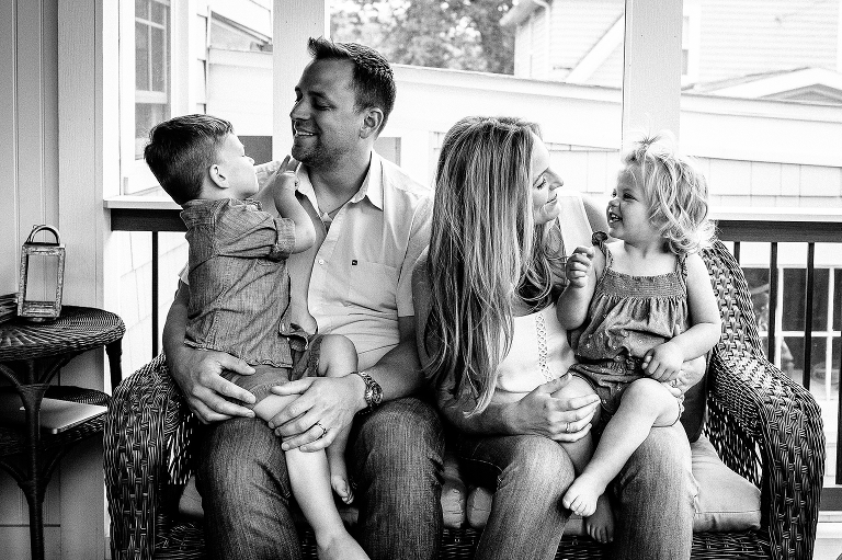 Black and white. Mother and father sit on chair on back porch with son and daughter.