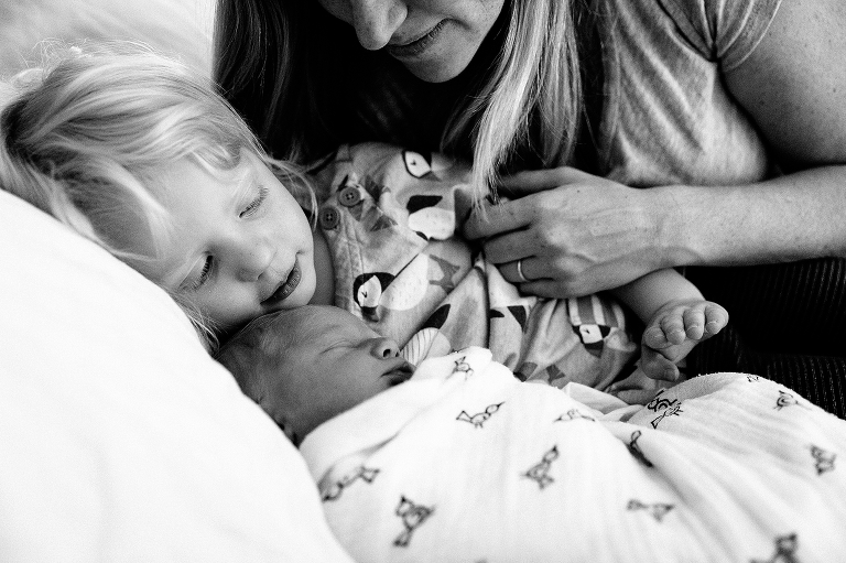 Black and white. Mother lays in bed with daughter hugging sleeping newborn baby 