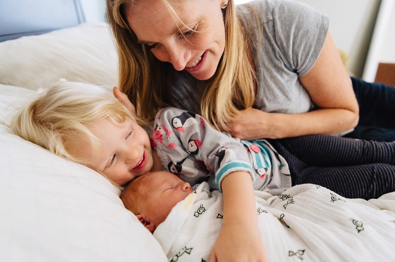 Mother lays in bed with daughter hugging sleeping newborn baby 