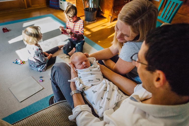 Mother and Father look at swaddled newborn baby while brother and sister play together on the floor