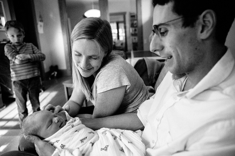 Black and white. Mom and Dad look at swaddled newborn baby with boy standing in the background.