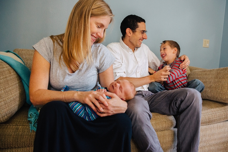Family sits on couch together. Mother holds newborn baby, father laughs with son