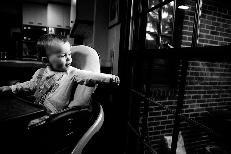 Black and white. baby sits in highchair looking and pointing out the window