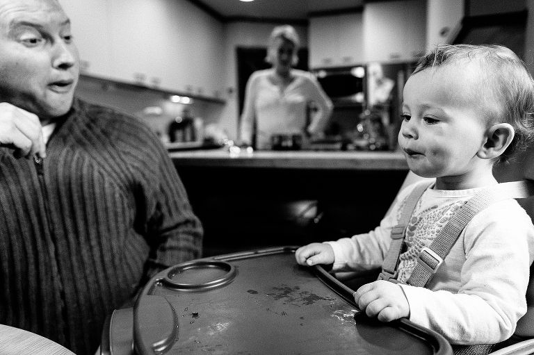 Black and white. Father sits with baby daughter at dinner table while mother watches from the kitchen