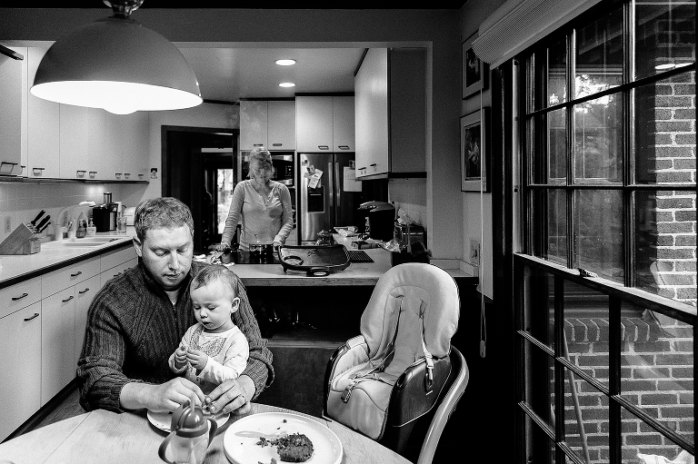Black and white, Daughter sits on fathers lap at the dinner table while mother cooks in the kitchen