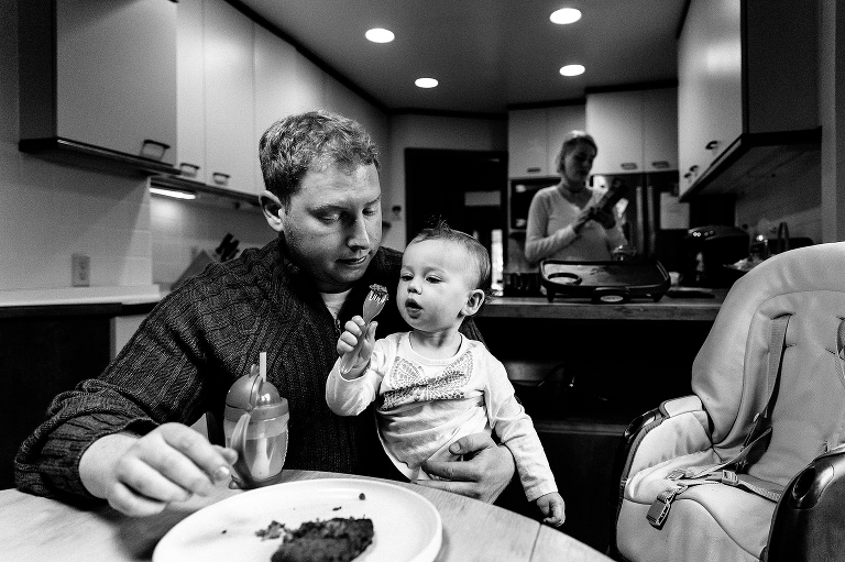 Black and white, Baby daughter sits on fathers lap while eating dinner, mother cooking in background