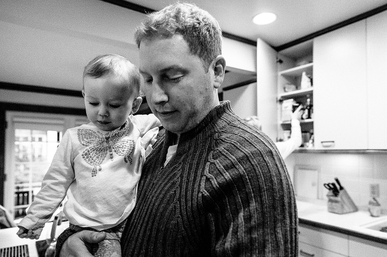 Black and whiteFather holds baby daughter in kitchen while mother looks in cabinet for dinner supplies