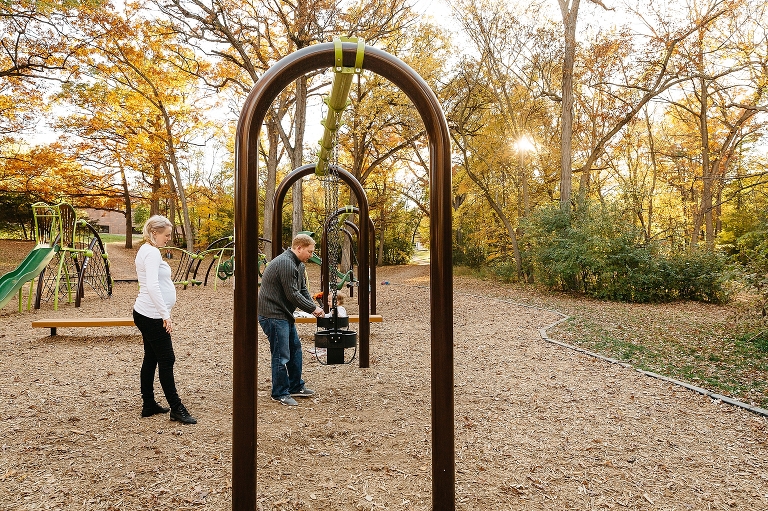 Father pushes baby in swing set at park while pregnant mom watches