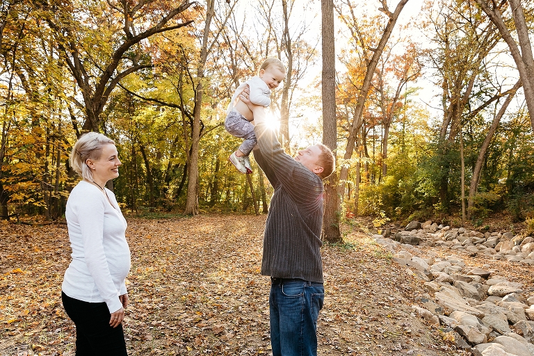 Father holds up baby with pregnant mom watching with sun shining in background in fall time