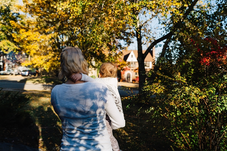 Mother holds baby in front yard at fall time with trees changing color