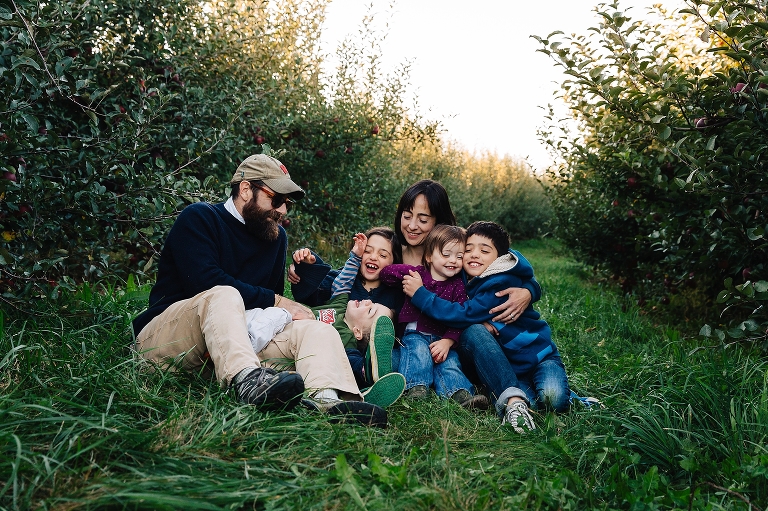 Mother, Father, and four kids sit in the grass at an apple orchard.