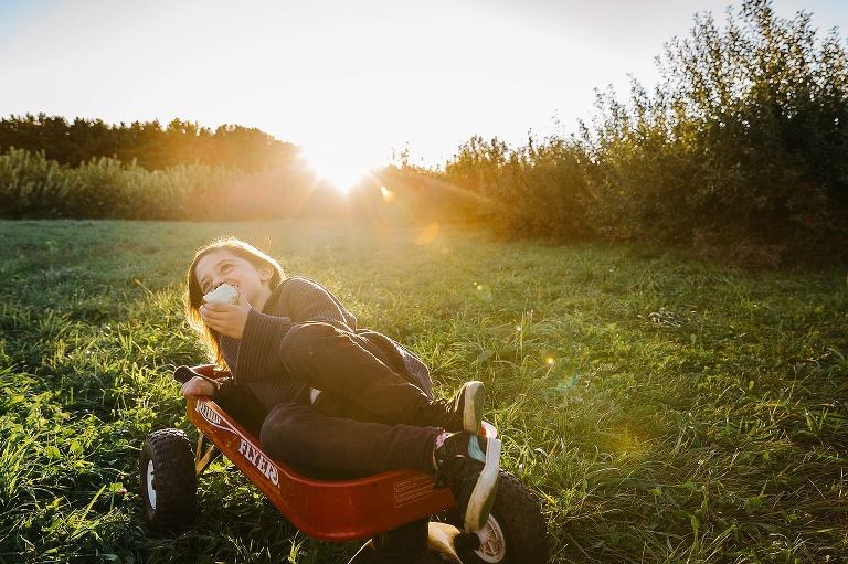 Girl sitting in red wagon in the grass eating an apple. Sun setting in the background shining light into camera.