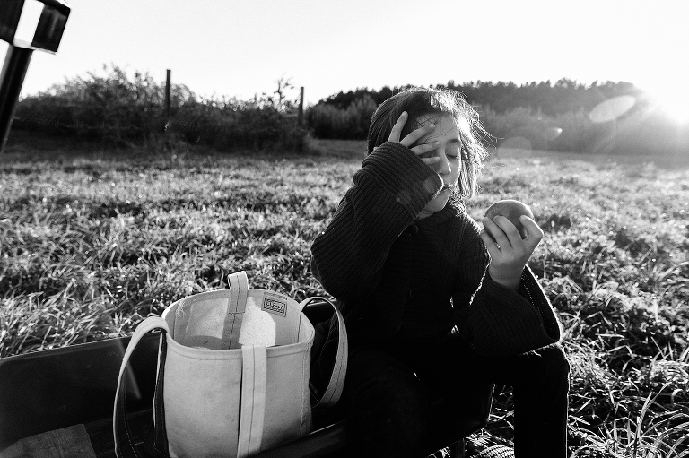 Black and white. Boy sits on grass eating an apple next to a wagon and canvas bag at a apple orchard.