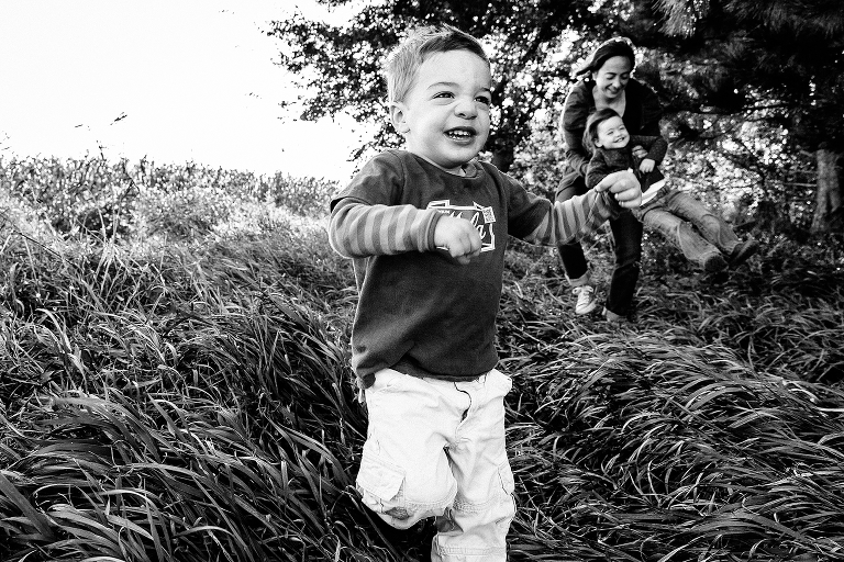 Black and white. Young boy runs down grassy path. mother picks up child in the background.