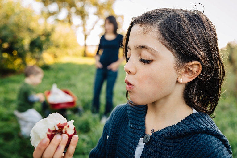Girl eats red apple at apple orchard. Mother and younger brother blurred in the background.