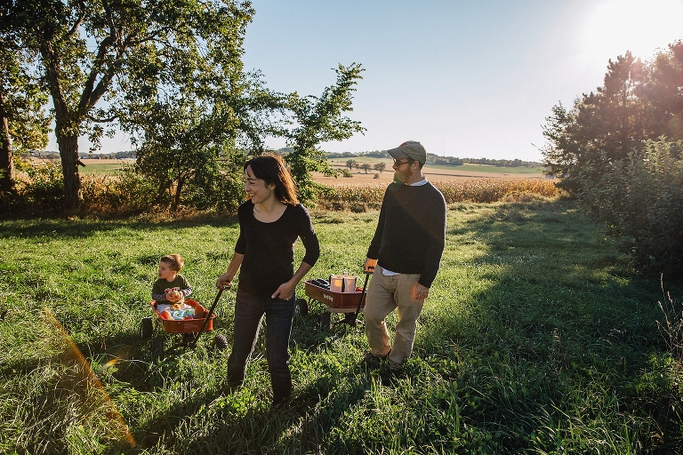 Location: local apple orchard. Father pulls red wagon full of fresh apples and mother pulls wagon with young son in it.