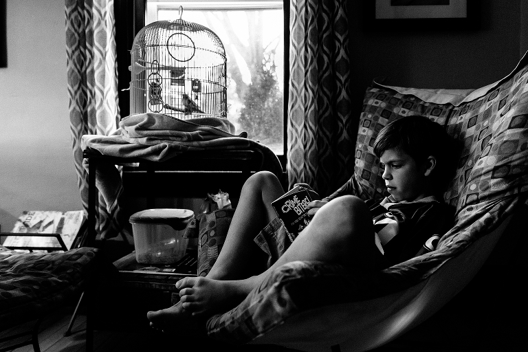 Black and white. Young boy sitting in chair next to bird cage reads book