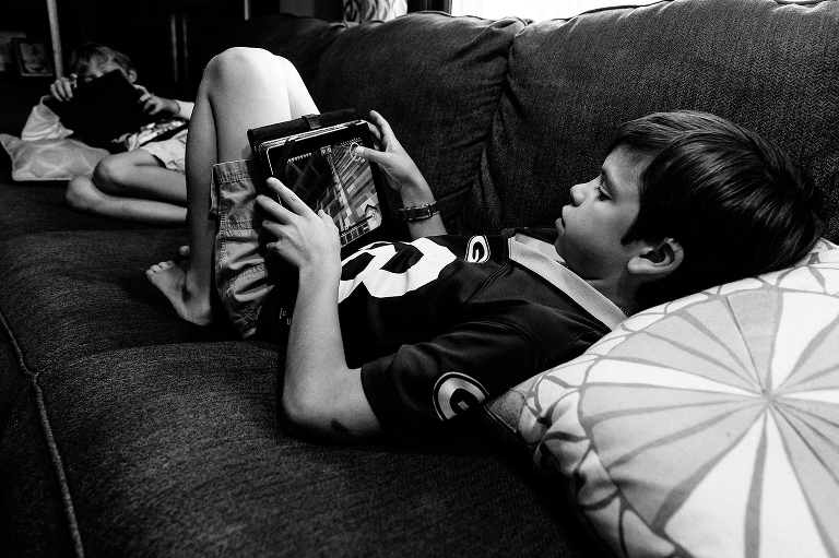 Black and white. Two brothers sit on couch both playing games on iPads