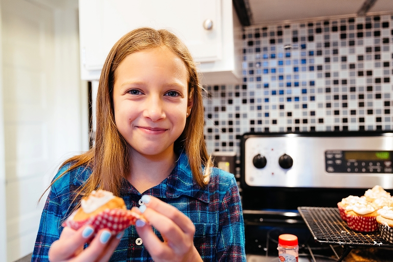 Girl stands in kitchen with freshly made cupcakes smiling and eating one