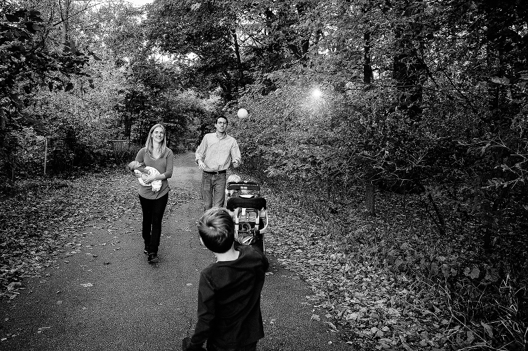 Black and white. Family of five walks down trail with trees all around them. Father and son play catch with baseball