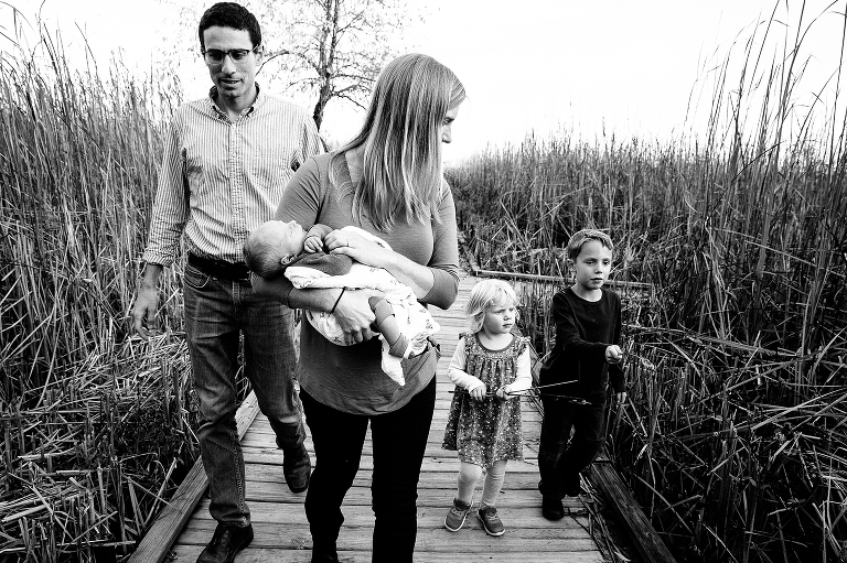 Black and white. Family of five walks down a boardwalk together, young boy and girl holding cattails.