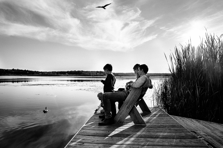 Black and white. Family sitting together on a dock bench look into the lake with duck swimming by and a bird flying above them