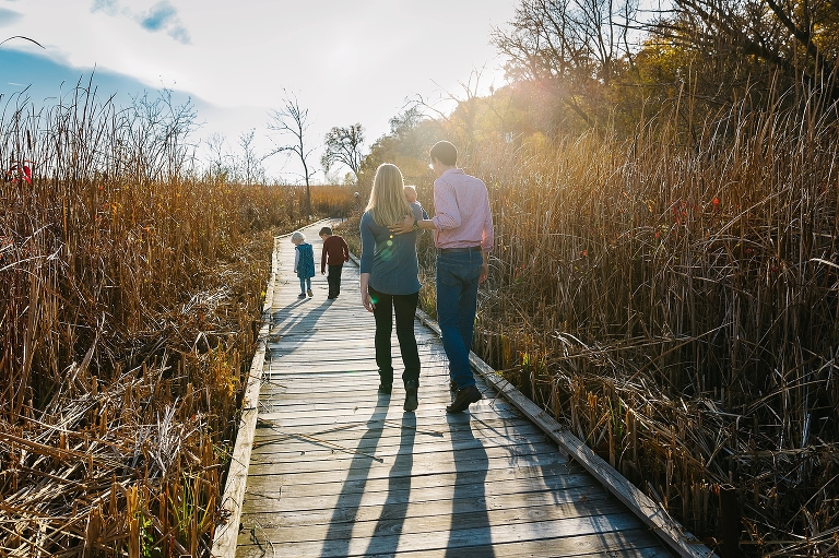 Family of five walks down a boardwalk together at sunset with cattails next to them