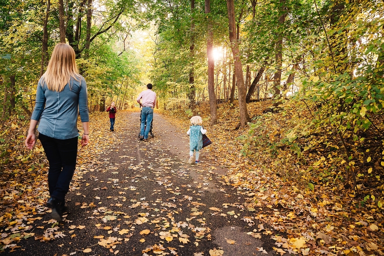 Family of five walks don path with colored tree leaves all around them with the sun setting through the trees