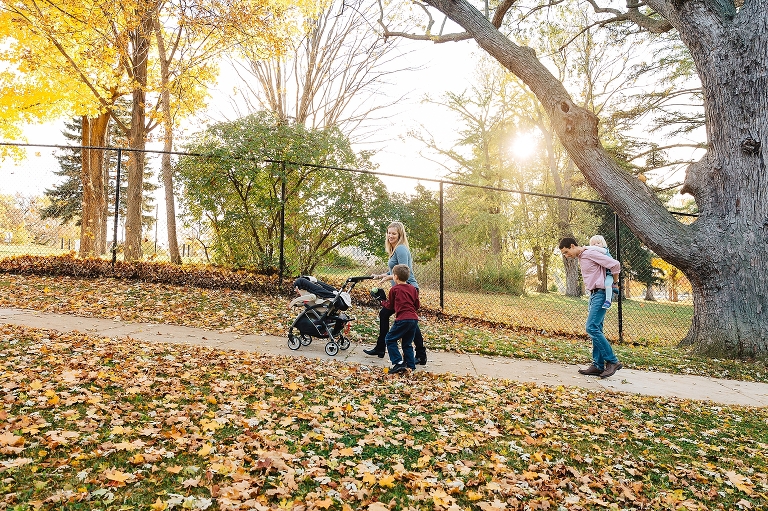 Family of five walks up hill together. mother pushing a stroller with newborn baby, son walking next to her, and father holding daughter on his back