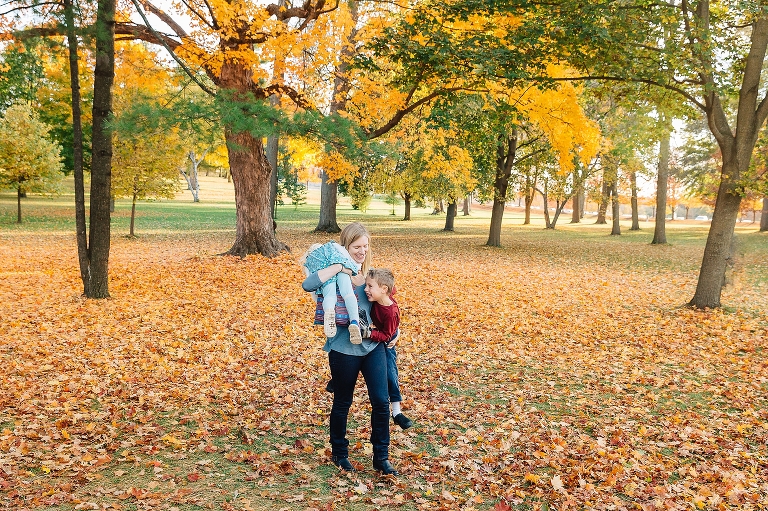 Mother carries laughing daughter and son at a park with red and orange leaves fallen all over the grass