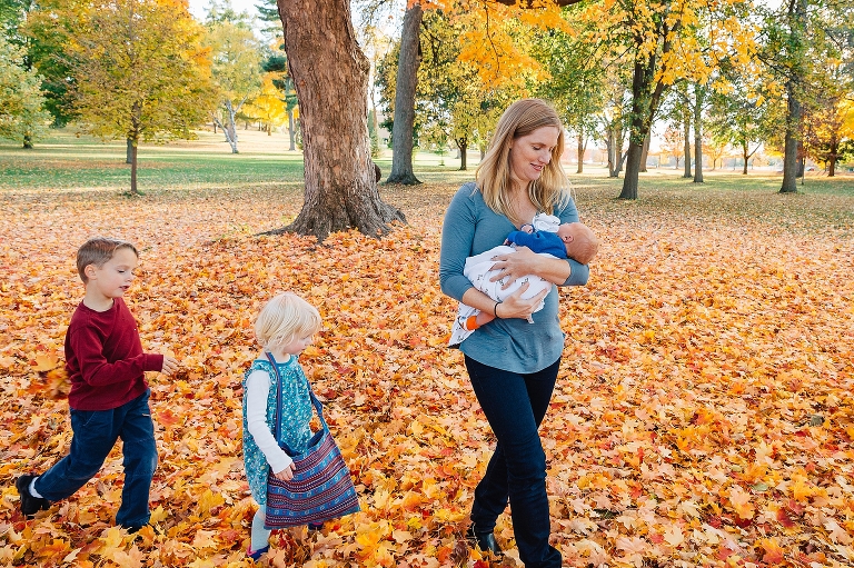 Mother holding newborn baby walks through a park at fall time with leaves all over the ground with son and daughter following behind her