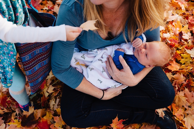 Mother sits in orange leaves holding newborn baby while older daughter hands mother a leaf