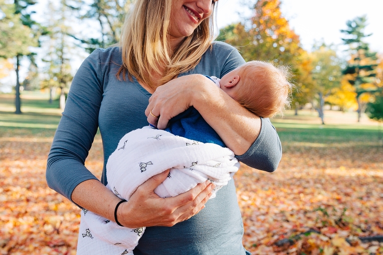 Mother holds newborn baby boy outside at a park with fallen red leaves all around them
