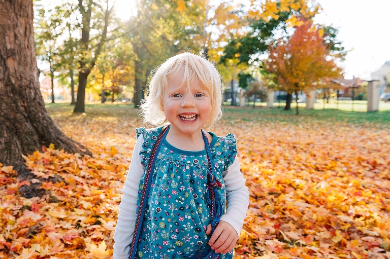 Toddler girl smiles outside at park standing in red fall leaves