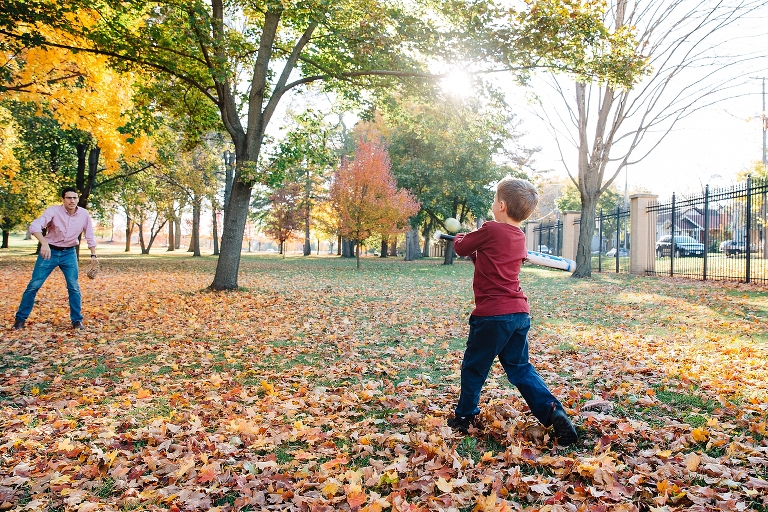 Dad plays baseball outside with his son at a park at fall time with colored leaves on the grass