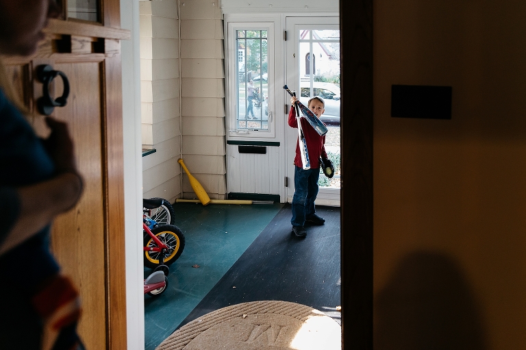 Boy walks outside of house carrying two baseball bats, mitt and a ball