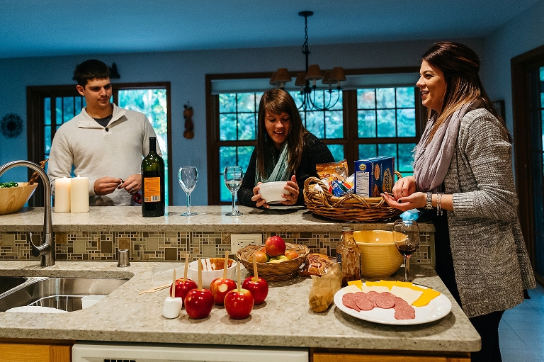 Family stands around kitchen with caramel apples, cheese and meat platter, wine, and smores.