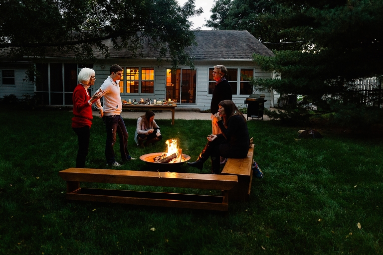 Family sits outside in backyard around fire pit eating smores.