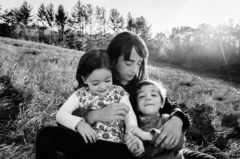Mother lays on a hillside with twin children. Sunflare comes over the trees. Black and white. 